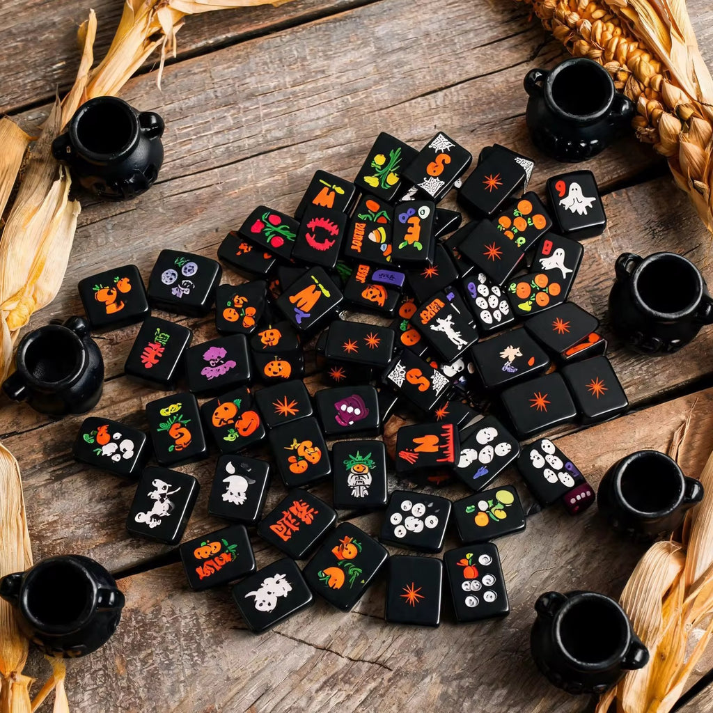 Black Halloween mahjong tiles with colorful ghost, pumpkin, skull and spooky designs scattered on a rustic wooden table.