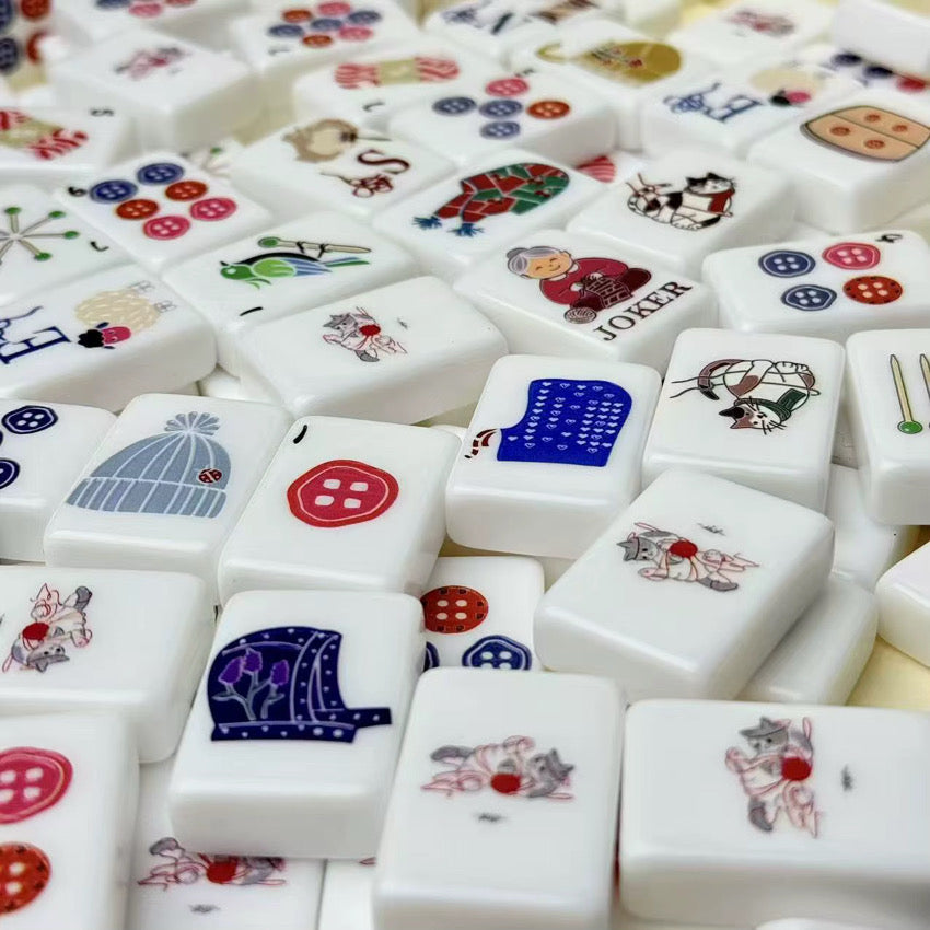 Close-up of scattered white mahjong tiles featuring knitting, yarn, cats, buttons, hats, and colorful craft designs
